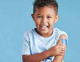 A smiling child points to a small bandage on his upper arm.